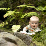 Puolivuotias vauva makaa vatsallaan viltin päällä metsässä saniaisten ympäröimänä. Vauvan suu on hieman auki ja pää ylhäällä, vauva kohottaa itseään käsillään.