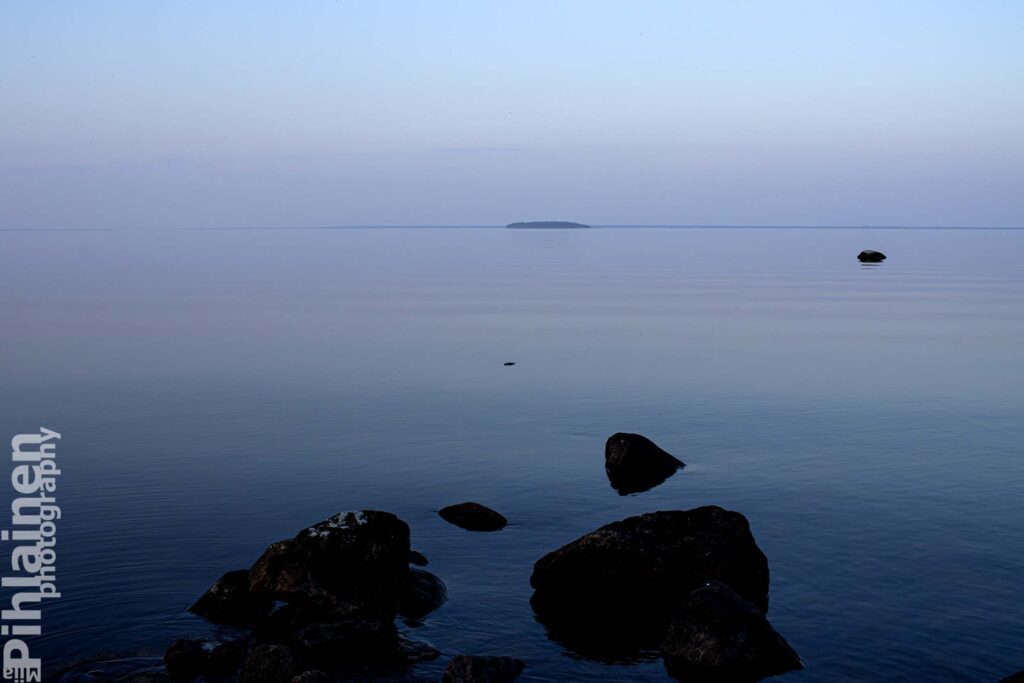 Oulujärvi on lähes peilityyni ilta-aikaan. Etualalla rannan kiviä ja horisontissa näkyy saari.