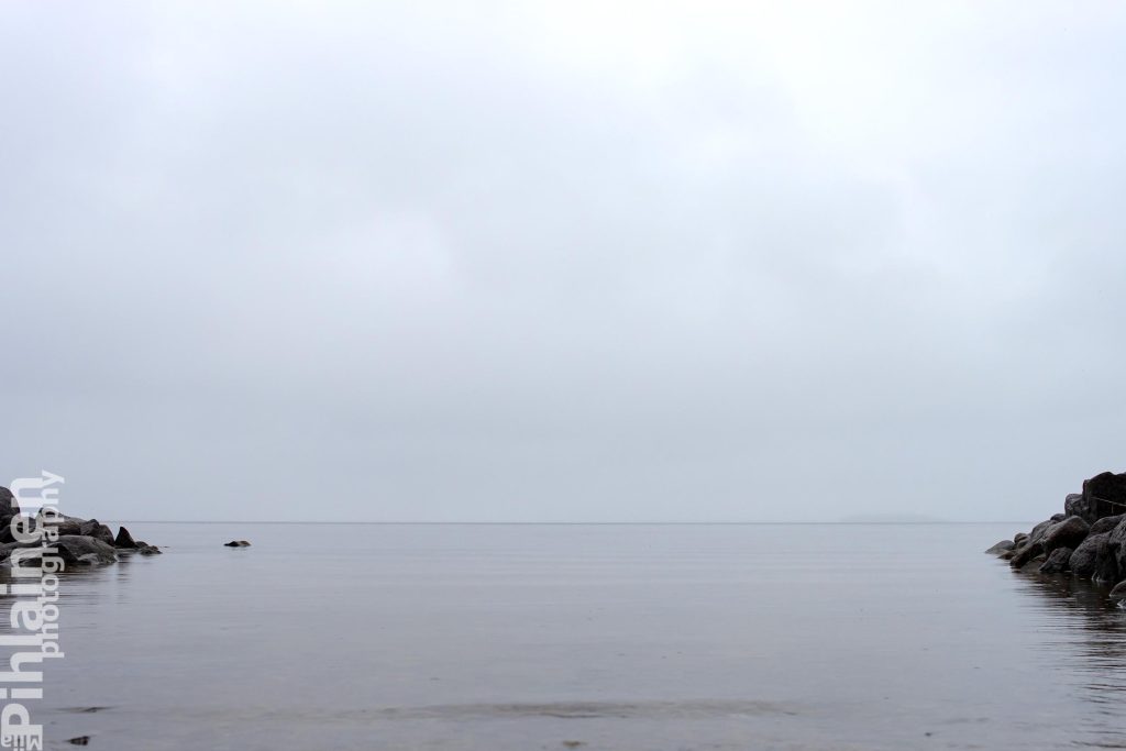 Oulujärven harmautta. Horisontti on sumea, koska järvellä sataa. Kuvan reunoissa näkyy hieman suuria rantakiviä.