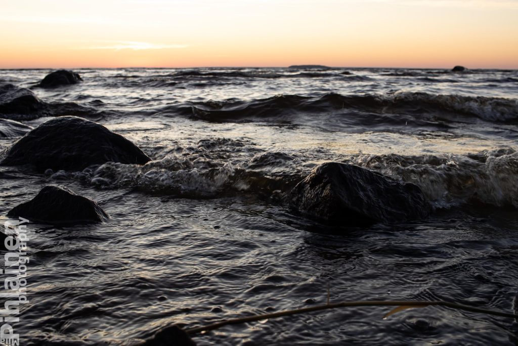 Oulujärvi auringonnousun aikaan: järvessa on aallokko ja etualalla rantakiviä, horisontti punertaa nousevan auringon merkiksi.